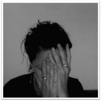 A person with dark hair covers their face with both hands, wearing rings on their fingers, sitting against a plain background. The image is in black and white.