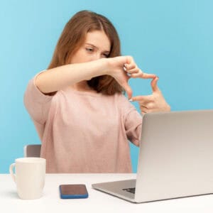 A young woman sits at a desk with a laptop, smartphone, and coffee mug, making a rectangular frame with her fingers, as if visualizing or focusing on something. The background is a solid light blue.