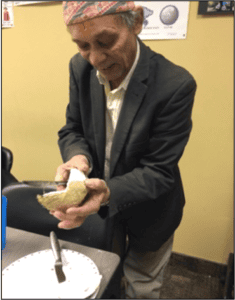 An elderly man wearing a traditional hat and suit jacket is holding and cutting a coconut over a table in an indoor setting. A plate and a knife are on the table in front of him.