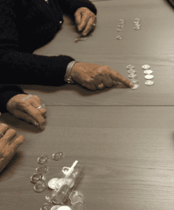 Three people sit at a table sorting and counting stacks of coins. One person points to a stack with their finger, while others have coins arranged in neat rows in front of them.