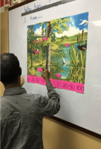 A person stands at a whiteboard placing pink number cards on a nature scene poster featuring a pond, trees, and birds. The board has "I have..." written on it above the poster.