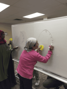 Two women draw large clock faces on a whiteboard using blue and red markers. Each holds a yellow teaching clock, and one woman wears a headscarf and pink sweater, while the other wears a green dress and red scarf.