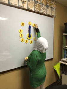 A person in a green outfit and white headscarf places yellow number magnets on a whiteboard arranged like a clock face, with a blue arrow as the clock’s hand. Educational posters are visible above the board.