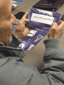 A person in a gray jacket holds a purple wrapped package with a label that reads, "How is the weather?" over a table with more similar packages.