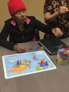 A woman wearing a red beanie places colorful game pieces on a world map on a table. Another person stands nearby, partially visible, holding a game piece. A container of pieces sits next to the map.