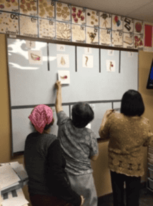 Three women are standing in front of a whiteboard with images and letters. One woman is reaching up to place a card on the board. Drawings of food and objects are displayed along the top border of the wall.