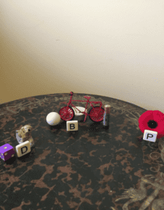 A small dog figurine, a purple die, a white ball, a red toy bicycle, a battery, and a red poppy flower are arranged on a round, dark-patterned table. Letter blocks "D," "B," and "P" are placed near the objects.