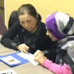 Two women sit at a table looking at documents together. One woman, in a dark coat, holds a pen and appears to be explaining something to the other, who is wearing a purple and gray headscarf and a pink sleeve.