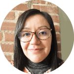 A woman with straight dark hair and glasses smiles at the camera. She is wearing a gray turtleneck and patterned sweater, standing in front of a brick wall background.