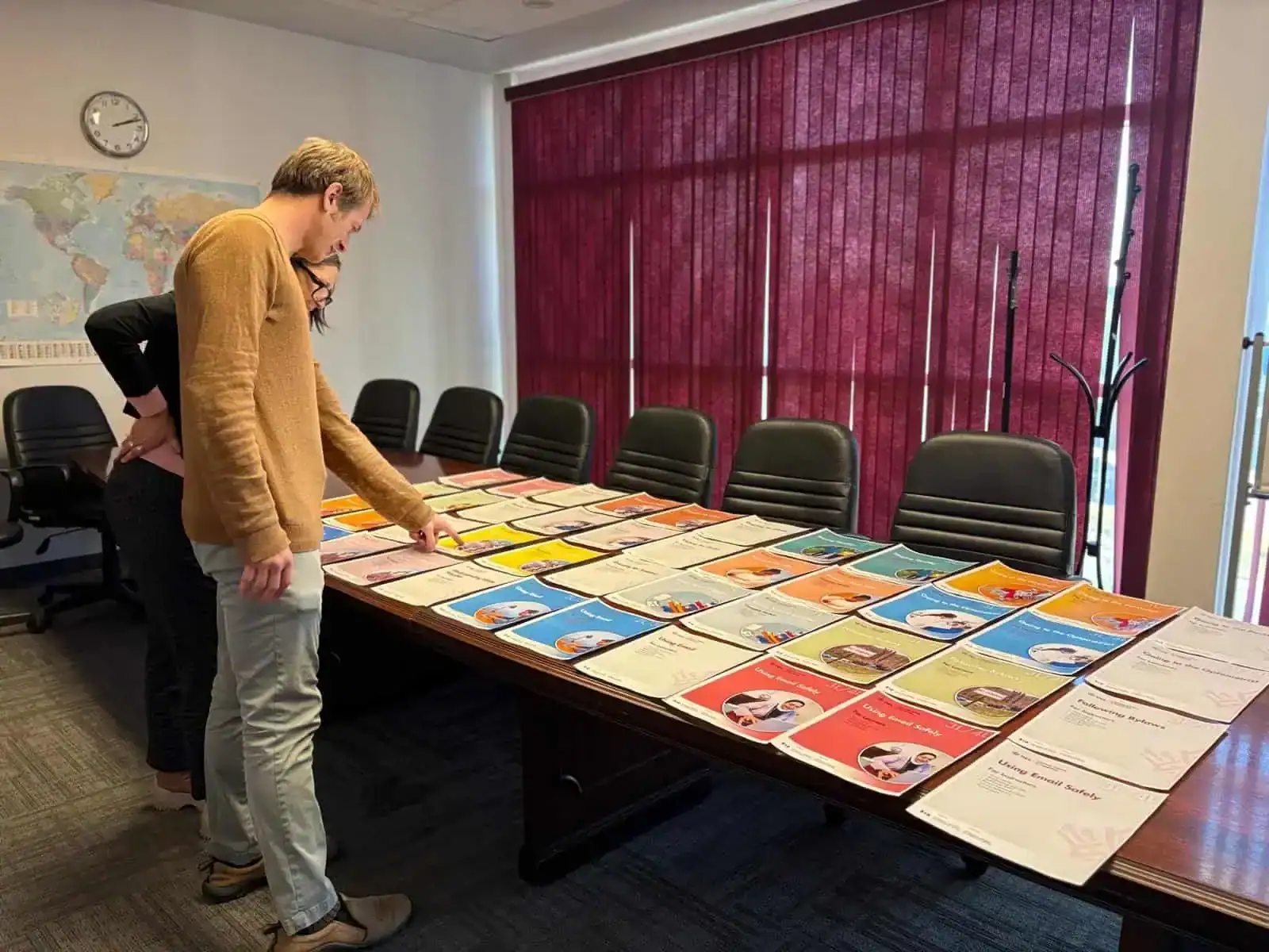 Two people stand by a long conference table covered with colorful certificates or booklets in the About Us meeting room, which features black chairs, a map on the wall, burgundy blinds, and a clock showing 10:10.