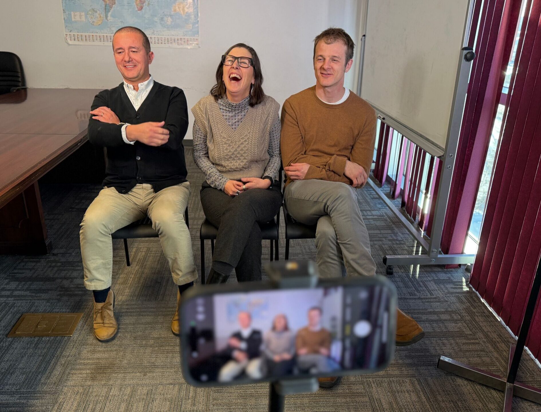 Three people sit side by side on chairs, smiling and laughing for an About Us video filmed by a smartphone on a tripod in an indoor office with a table, map, and red vertical blinds in the background.