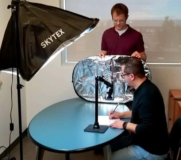 Two people set up a photography shoot at a table for an "About Us" feature. One adjusts a light and holds a reflective panel, while the other writes on paper under a mounted camera. Professional lighting equipment is visible.