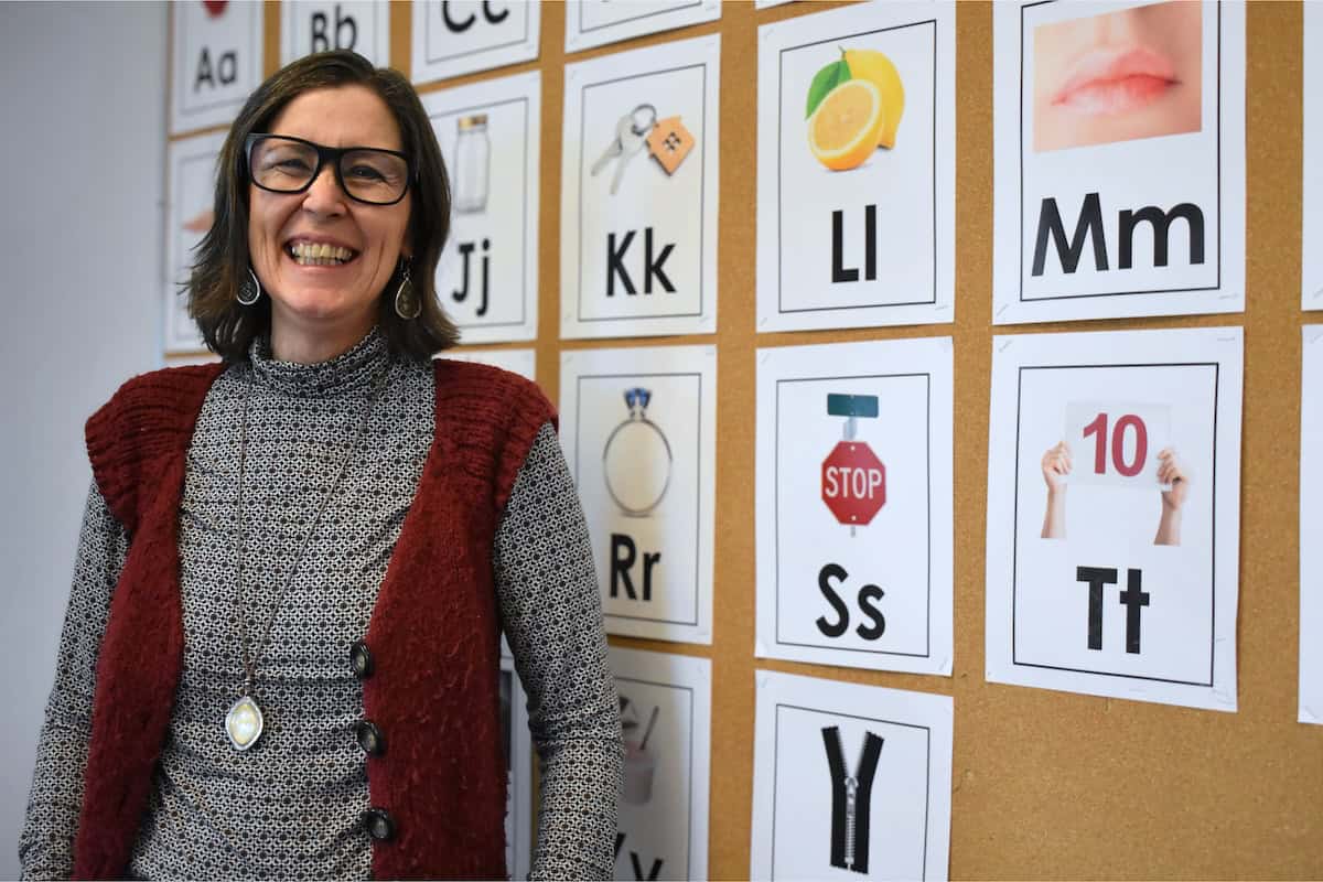 A smiling woman wearing glasses and a red vest stands in front of an "About Us" wall with alphabet cards, each displaying a letter and corresponding image, such as a lemon for "Ll" and a stop sign for "Ss.