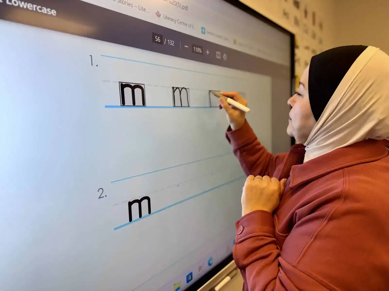 A woman wearing a headscarf and red jacket writes the lowercase letter “m” on an interactive whiteboard at the Literacy Centre of Expertise, practicing penmanship along dotted lines.