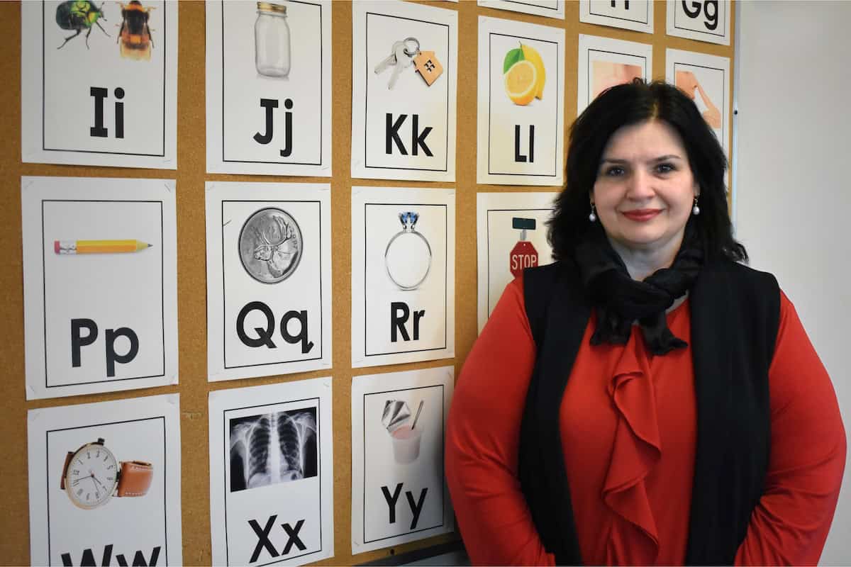 A woman in a red blouse stands smiling in front of a bulletin board with alphabet cards for our About Us page, each displaying a letter and an image, such as a pencil for P and a lemon for L.