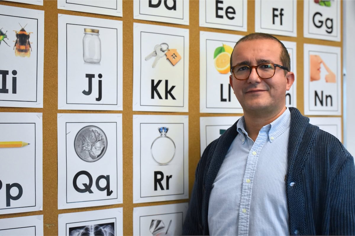 About Us: A man wearing glasses and a blue sweater stands in front of a wall with alphabet cards, each featuring a letter and a corresponding picture, such as a jar for "Jj" and keys for "Kk.